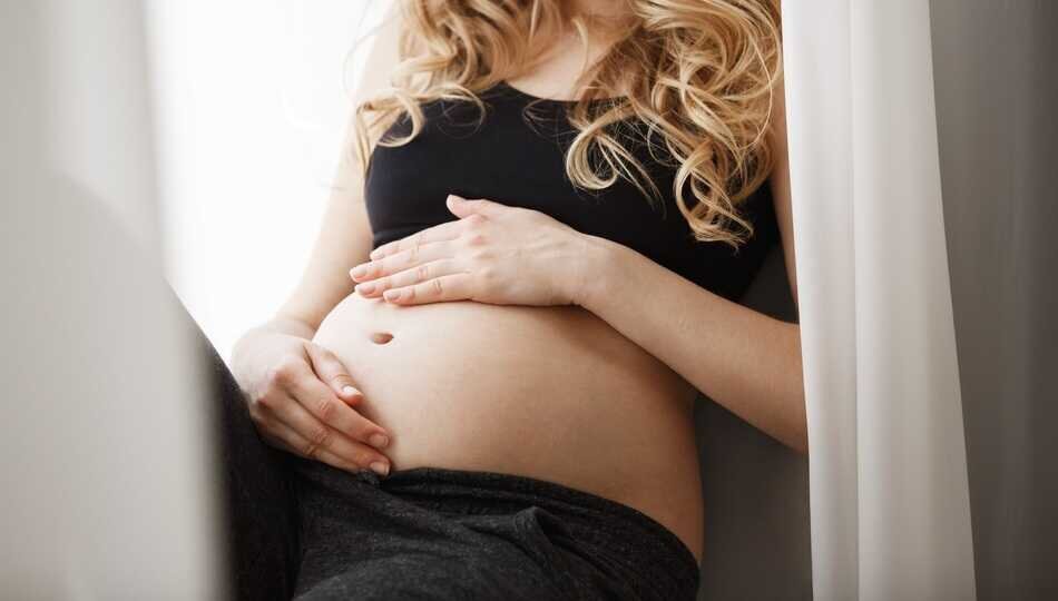 Close up detail of young attractive blonde pregnant mother in black outfit sitting on window sill in bedroom, touching and looking at belly with happy expression. Maternity concept.